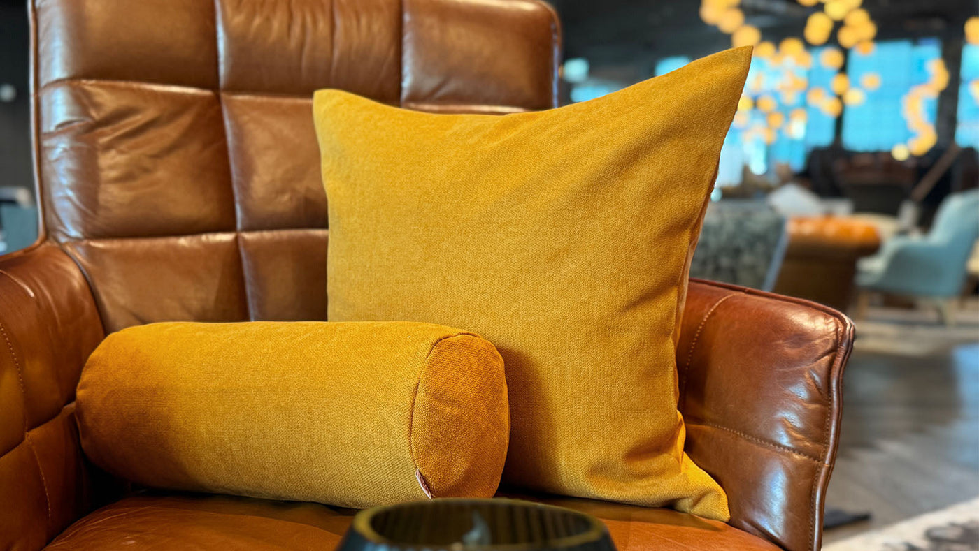 Ein senfgelbes quadratisches Kissen und ein passendes Nackenkissen liegen auf einem braunen Ledersessel. Eine schwarze gerippte Vase steht im Vordergrund, und warmes Licht erhellt sanft den Hintergrund.
