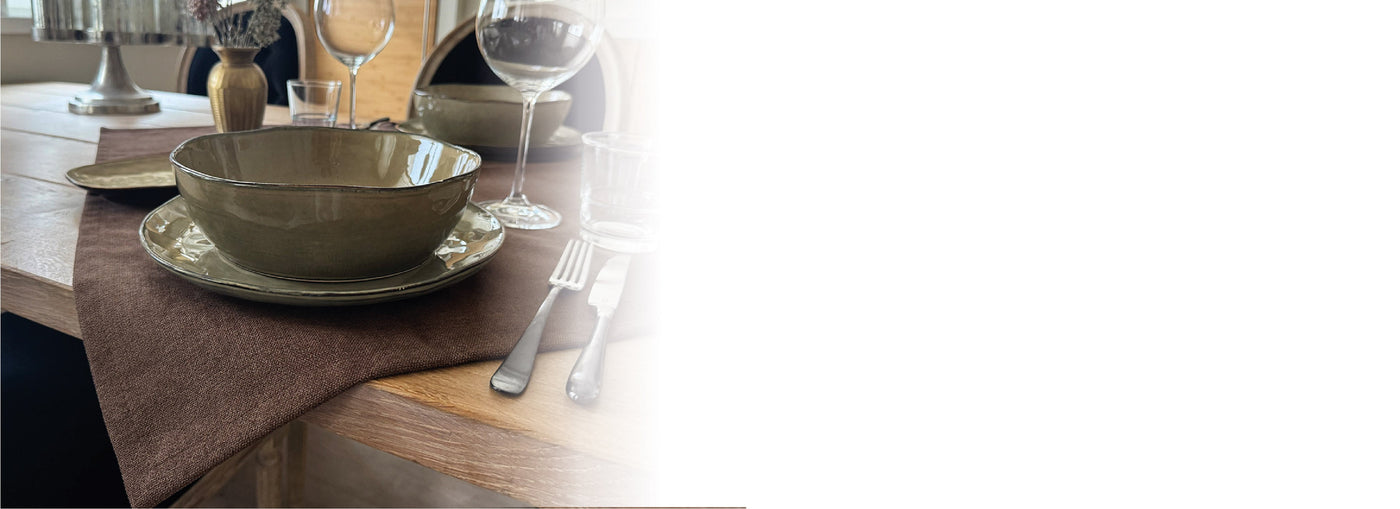 Nahaufnahme eines gedeckten Tisches mit grüner Keramikschüssel, Teller, Gabel, Messer und Weinglas auf einer braunen Tischdecke.