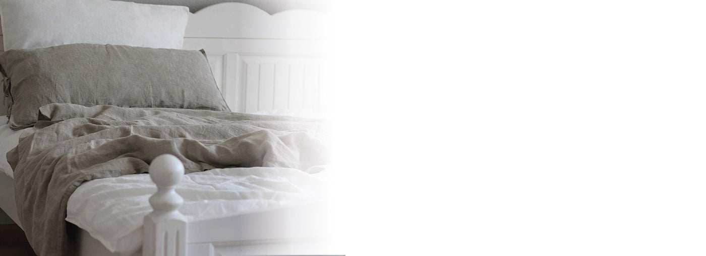Ein fein säuberlich gemachtes Bett mit weißen Kissen und Laken, darüber eine hellgraue Leinenbettdecke mit passendem Kissenbezug, in einem sanft beleuchteten, gemütlichen Schlafzimmer. Die linke Seite des Bildes verschwindet in Weiß.