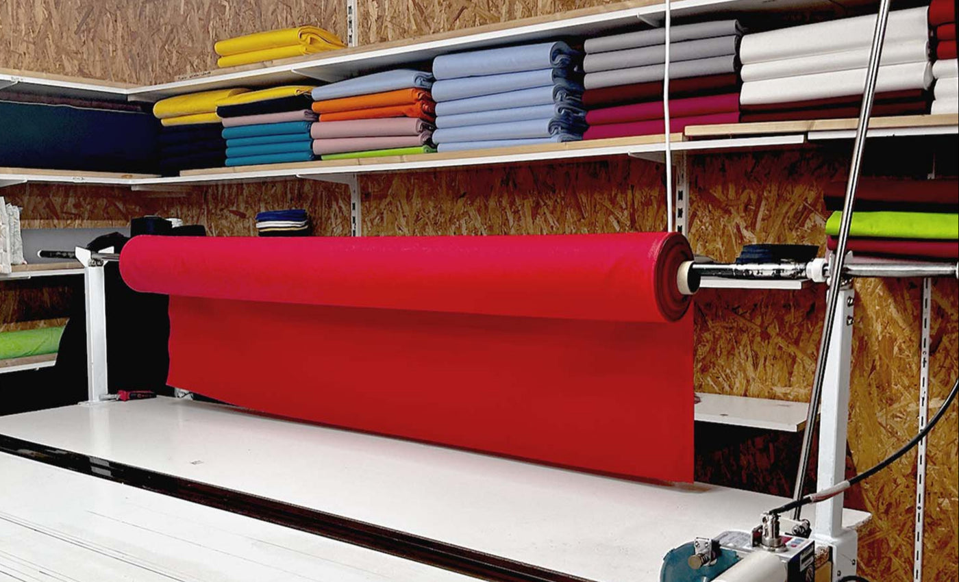 Eine große rote Stoffrolle liegt auf einem Zuschneidetisch. Im Hintergrund sind Regale mit ordentlich gestapelten, bunten Stoffrollen.