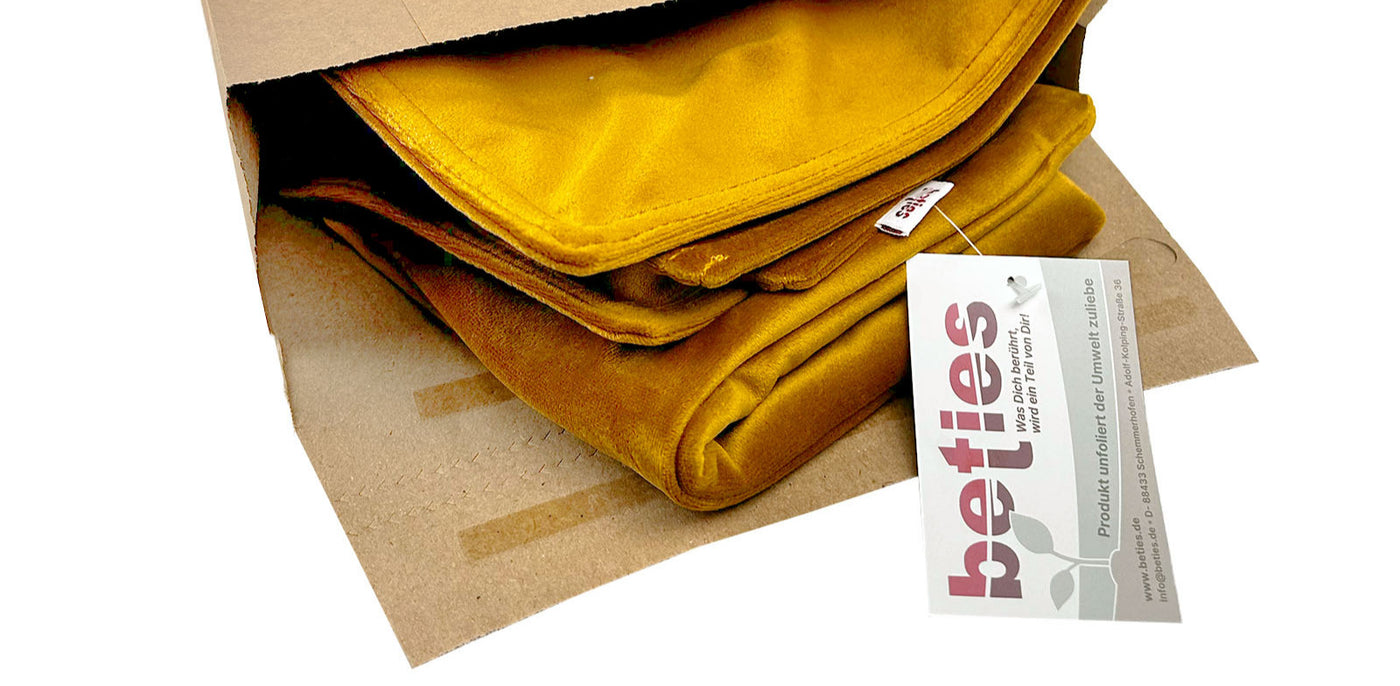 Ein senfgelber Kissenbezug , der teilweise aus einer braunen Papiertüte herausschaut. An dem Kissenbezug ist ein weißes Produktetikett mit dem Branding und Text von betties angebracht.