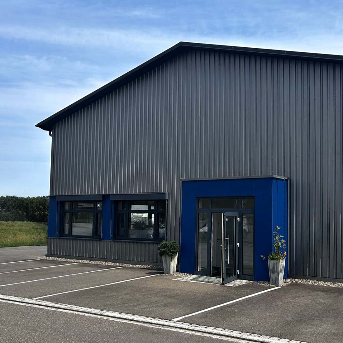 Ein modernes Industriegebäude mit grauen Metallverkleidungen, blauen Akzenten am Eingang, Glastüren, zwei großen Fenstern und Pflanzkübeln mit kleinen Bäumen vor einem teilweise bewölkten Himmel.