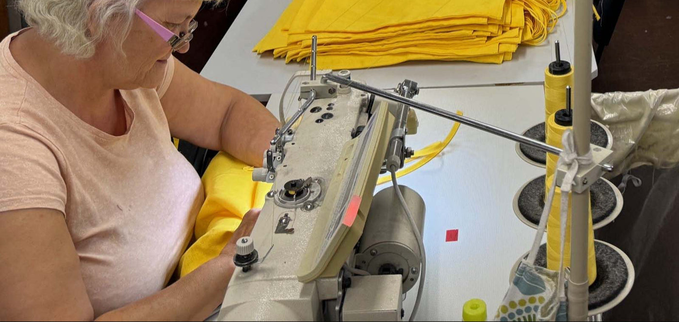 Eine Person mit weißem Haar und Brille näht mit einer Industrienähmaschine leuchtend gelben Stoff. Auf dem Tisch daneben liegen Stapel von gelbem Stoff und große Spulen mit gelbem Garn.