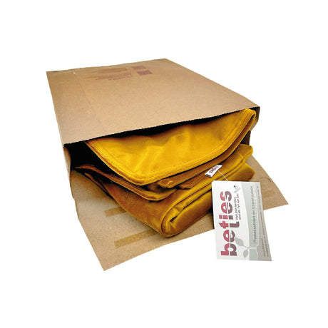 Ein senfgelber Kissenbezug , der teilweise aus einer braunen Papiertüte herausschaut. An dem Kissenbezug ist ein weißes Produktetikett mit dem Branding und Text von betties angebracht, auf weißem Hintergrund.