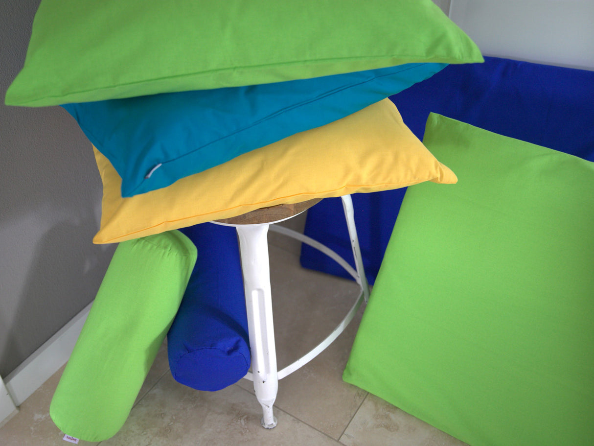 Hocker umgeben mit quadratischen Kissen, Nackenrollen und Keilkissen in Kissenbezuegen in den Unifarben Gelb, Petrol, Royalblau und Gruen.