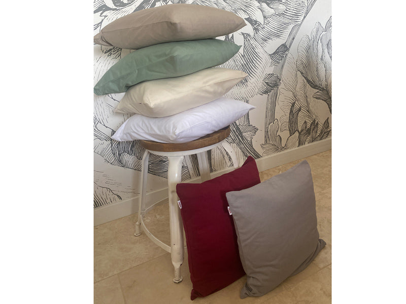 Hocker mit einem Stapel quadratischer Kissen bezogen mit Kissenbezuegen in den Uni-Farben Weiss, Natur, Gruen und Beige. Davor lehnen Kissen mit Bezuegen in den Farben Weinrot und Grau.