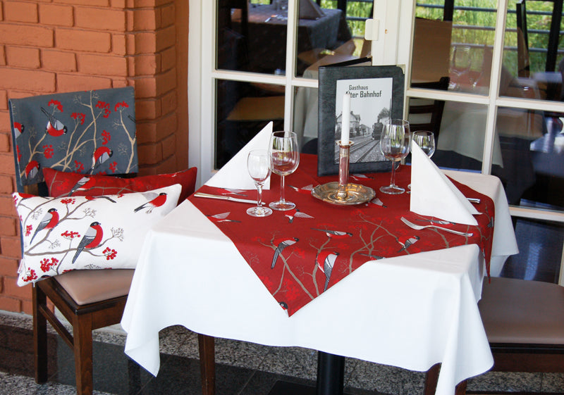 Rote Mitteldecke mit rot-grau-schwarzen Voegeln auf einem Tisch. Teilweise fliegend und teilwiese sitzen die Voegel auf Zweigen mit roten Beeren.