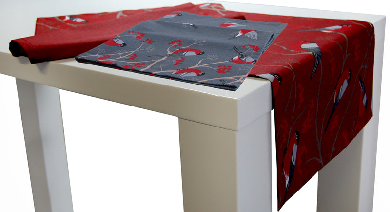 Grauer und Roter Tischlaeufer auf einem weissen Tisch. Das Motiv auf der Tischwaesche, sitzende und fliegende rot-grau-schwarze Voegel auf Zweigen mit roten Beeren.