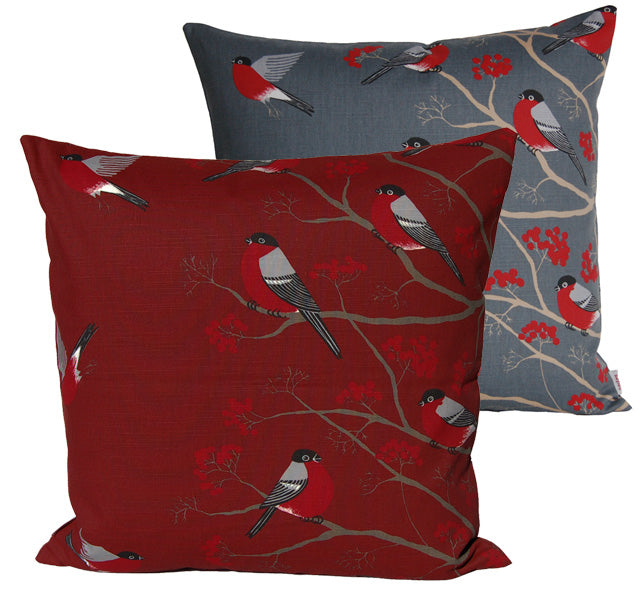 Ein rotes und ein graues quadratisches Kissen auf einem weissen Hintergrund. Die Kissenbezuege sind mit rot-grau-schwarzen Voegeln, die auf Zweigen mit roten Beeren sitzen, bedruckt.