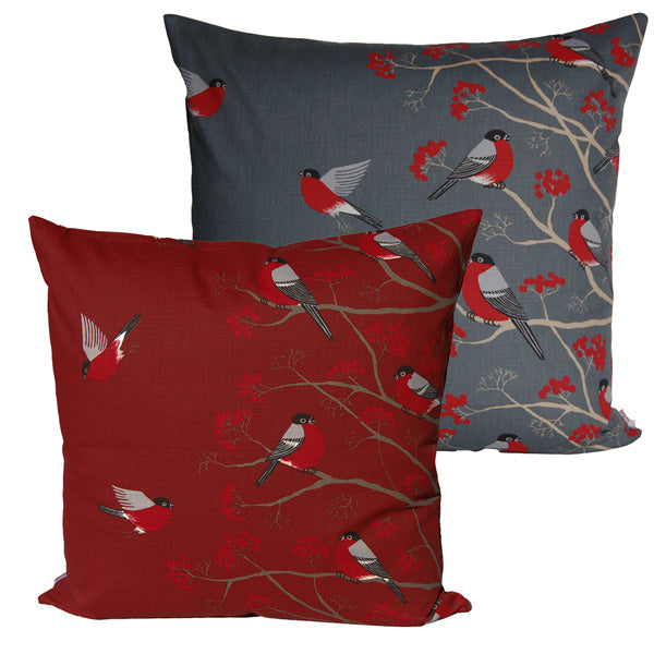 Ein rotes und ein graues quadratisches Kissen auf einem weissen Hintergrund. Die Kissenbezuege sind mit rot-grau-schwarzen Voegeln, die auf Zweigen mit roten Beeren sitzen, bedruckt.