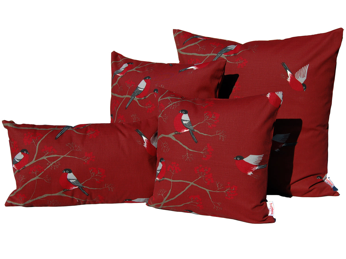 Drei quadratische und ein rechteckiges rotes Kissen in verschiedenen Groessen auf einem weissen Hintergrund. Die Kissenbezuege sind bedruckt mit rot-grau-schwarzen Voegeln die auf Zweigen mit roten Beeren sitzen oder teilweise fliegen.
