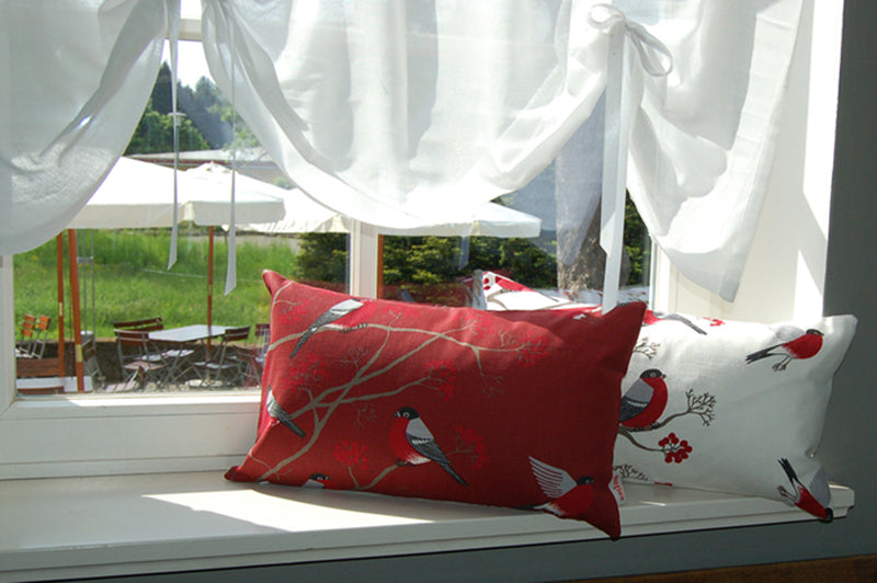 Fenster mit zwei Kissen auf der Fensterbank. Ein Kissen ist mit einem roten und das zweite Kissen mit einem weissen Bezug bezogen. Das Motiv auf den Kissenbezuegen, sitzende rot-grau-schwarze Voegel auf Zweigen mit roten Beeren.