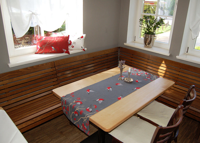 Grauer Tischlaeufer mit rot-grau-schwarzen Voegeln auf einem hellen Holztisch. Auf der Fensterbak liegen zwei Kissen, ein rotes und ein weisses, mit dem gleichen Motiv.