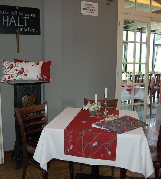 Roter Tischlaeufer mit rot-grau-schwarzen Voegeln auf einem Tisch. Teilweise fliegend und teilwiese sitzen die Voegel auf Zweigen mit roten Beeren.