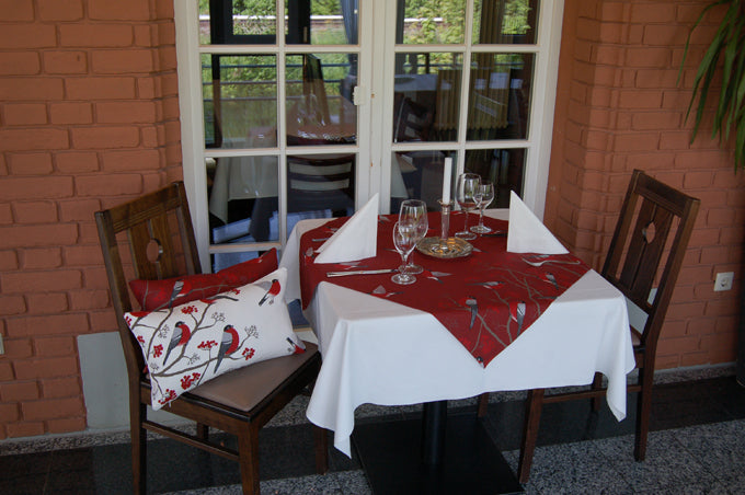 Rote Mitteldecke mit rot-grau-schwarzen Voegeln auf einem Tisch. Teilweise fliegend und teilwiese sitzen die Voegel auf Zweigen mit roten Beeren.
