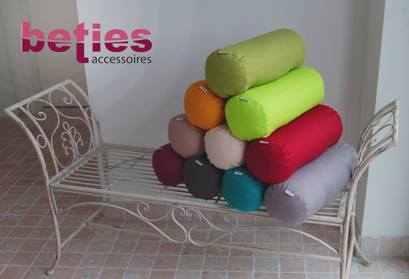 Nackenrollen als Pyramide auf einer weissen Eisenbank gestapelt. Bezogen mit einfarbigen Kissenbezuegen in den Farben Hibiskus-Beere, Schiefer-Grau, Petrol, Platin-Grau, Toffee, Natur-Beige, Karmin-Rot, Gold-Gelb, Apfel-Gruen und Gruen.