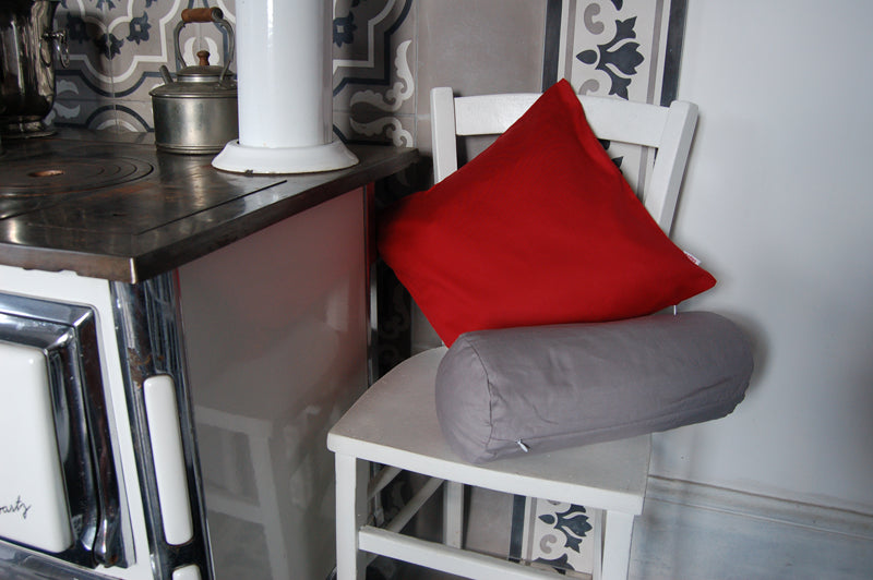Neben einem alten Holz-Kuechenofen steht ein weisser Kuechenstuhl. Darauf ist ein rotes und ein graues Kissen dekoriert.