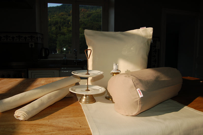 Ein Hellbeiges Kissen und ein hellbeiger Tischl�ufer mit einer beigen Nackenrolle auf einem Tisch drapiert.