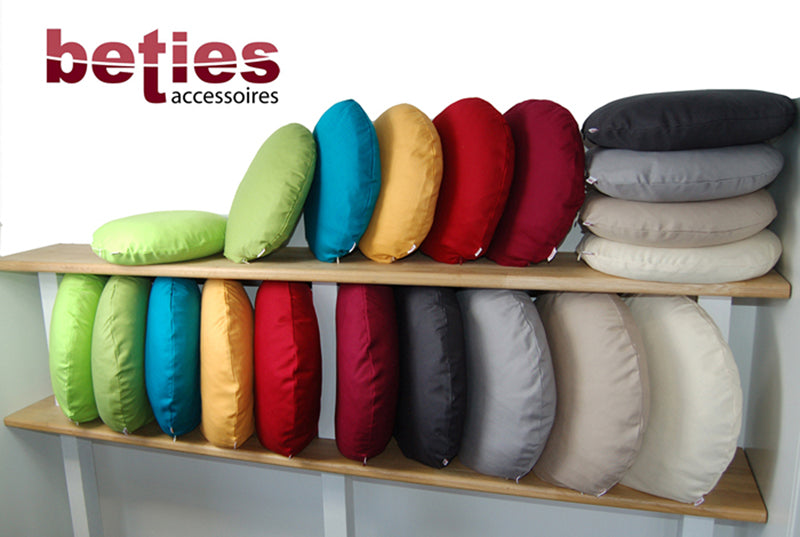 Zwei Reihen Runde Kissen in einem Holzregal. Die Kissen sind mit einfarbigen Kissenhuellen, in den Farben bezogen.