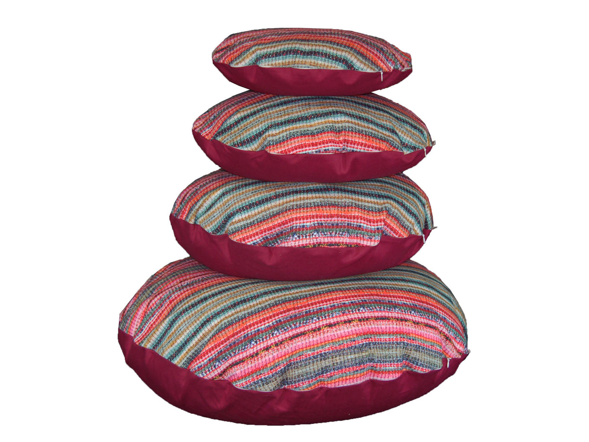 Gestreifte, runde Wende-Kissenbezuege, gestapelt als Turm. Die Vorderseiten sind gestreift in verschiedenen rot, rosa, gruen und blau Tönen. Die Rueckseiten sind uni hibiskus-weinrot.