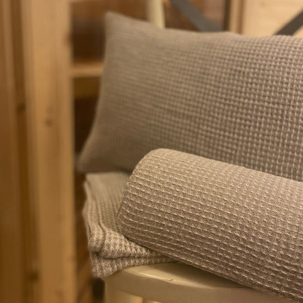 Auf einem Hocker liegt ein Saunatuch, eine Halb-Nackenrolle und ein Kissen, mit einem Bezug aus beigem Waffelpique-Stoff bezogen.