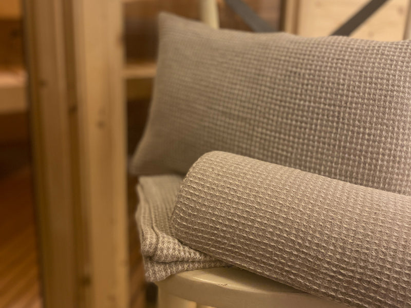 Auf einem Hocker liegt ein Saunatuch, eine Halb-Nackenrolle und ein Kissen, mit einem Bezug aus beigem Waffelpique-Stoff bezogen.