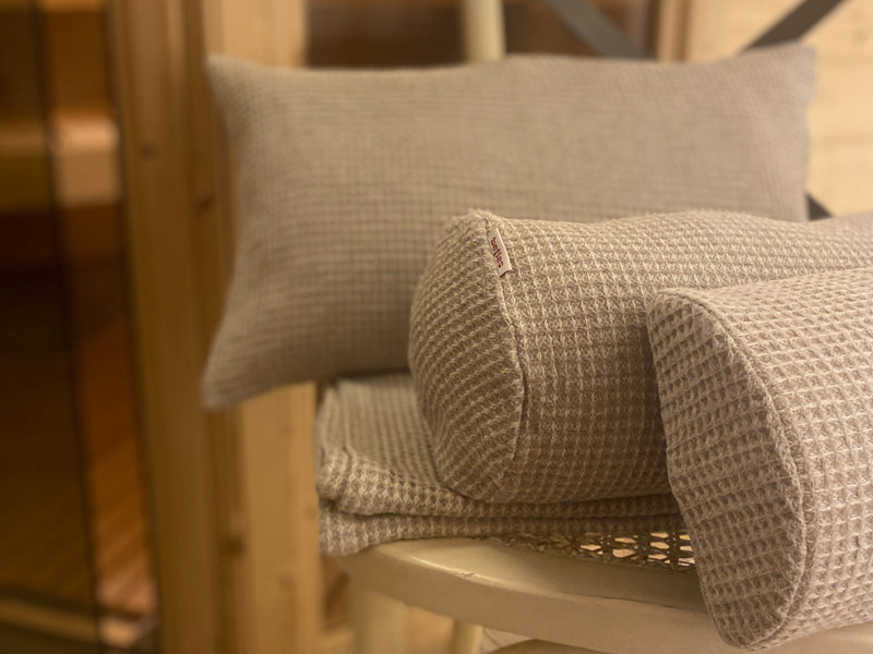 Auf einem Saunatuch aus beigem Waffelpique-Stoff liegt eine Halb-Nackenrolle, eine Nackenrolle und ein Kissen. Alle drei sind mit einem Bezug aus beigem Waffelpique-Stoff bezogen.