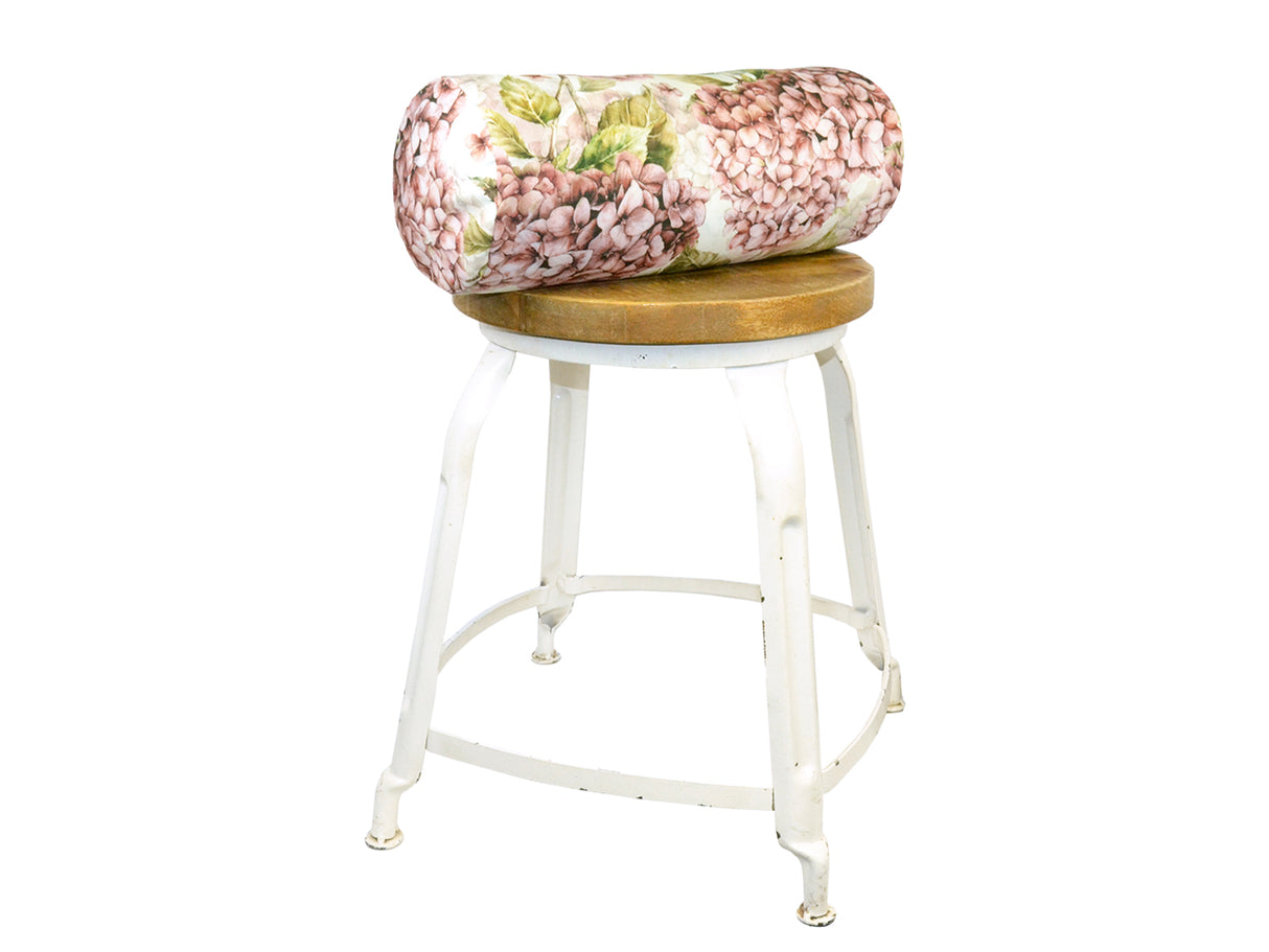 Auf einem Hocker liegt ein weisser Bezug für Nackenrollen im Landhausstil, bedruckt mit rosa/pinken Hortensien-Blueten.