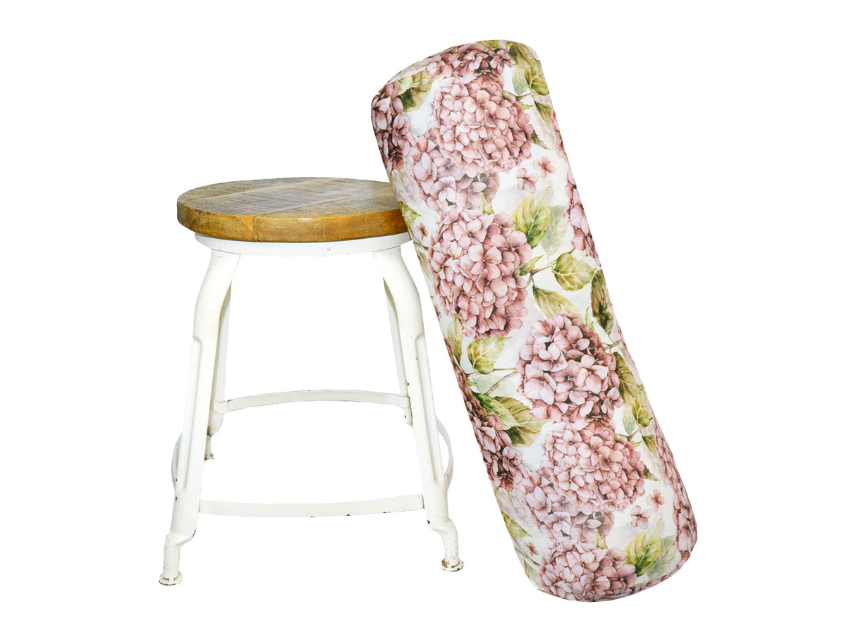 An einem Hocker lehnt ein XL-Rollen-Kissen bezogen mit einem Bezug im Landhausstil, bedruckt mit rosa/pinken Hortensien-Blueten.