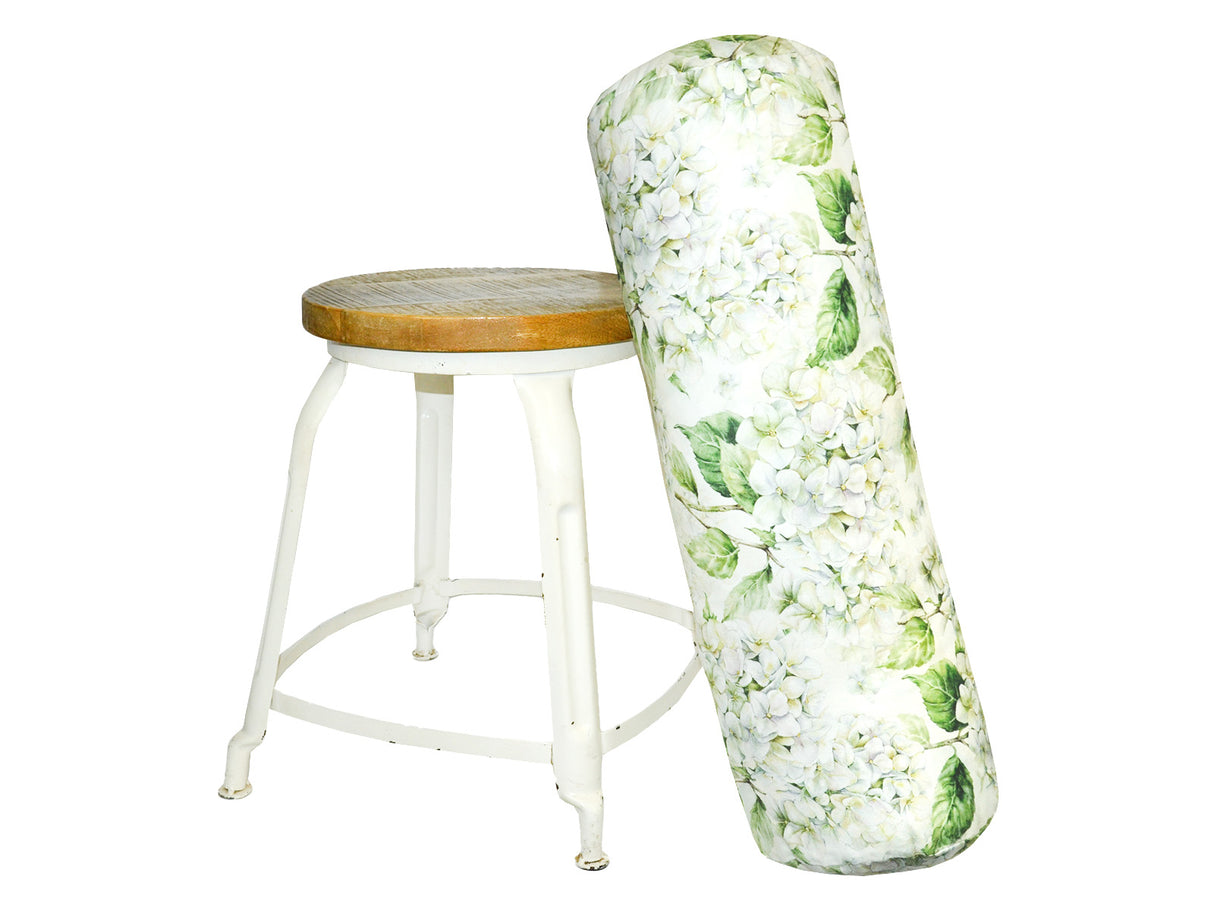 An einem Hocker lehnt ein XL-Rollen-Kissen bezogen mit einem Bezug im Landhausstil, bedruckt mit rosa/pinken Hortensien-Blueten.