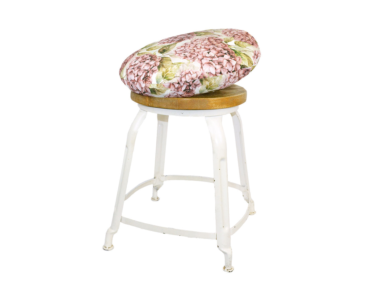Auf einem Hocker liegt ein runder Kissenbezug im Landhausstil, bedruckt mit rosanen/pinken Hortensien-Blueten.