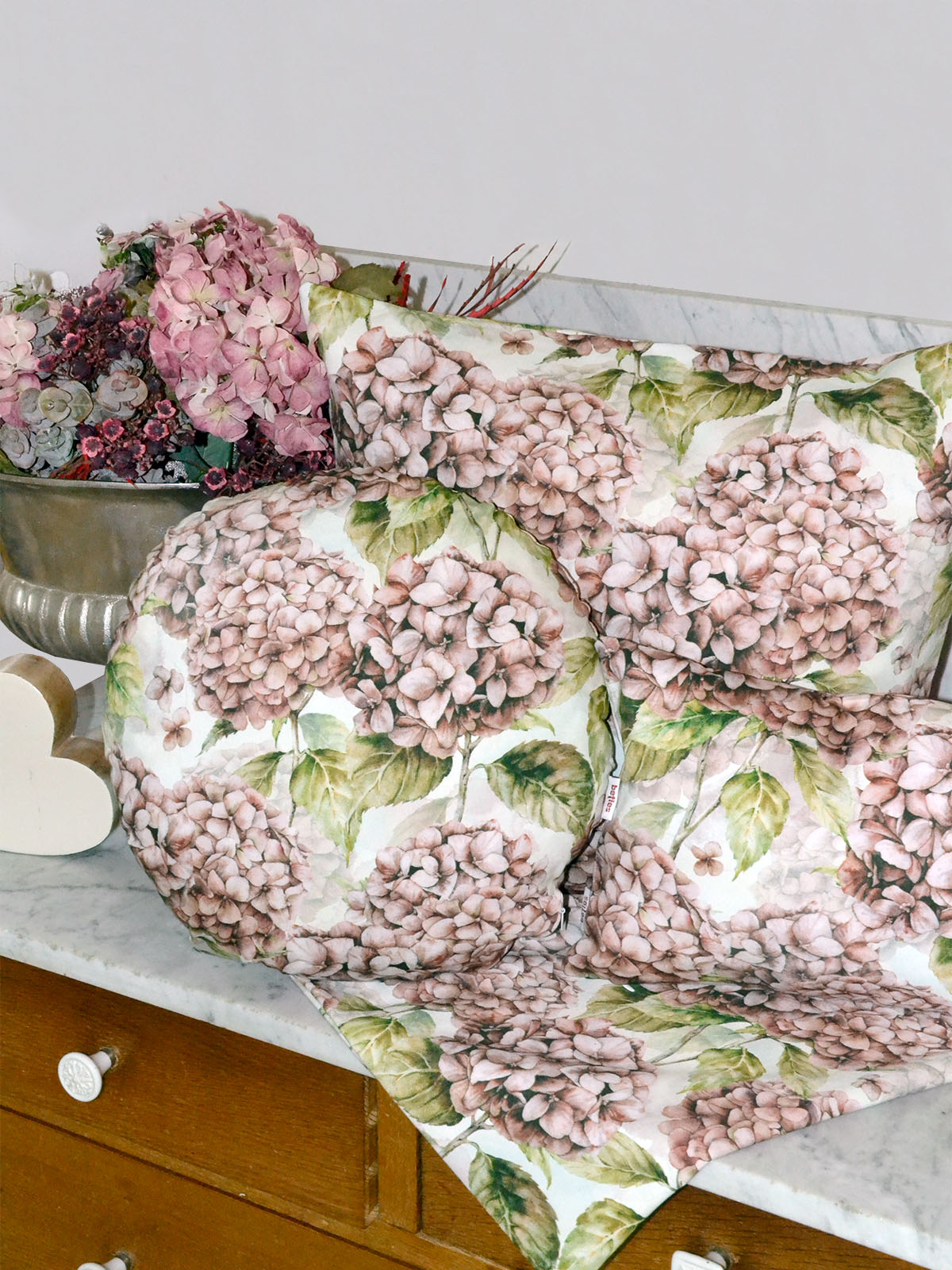 Drei Kissen bezogen mit je einem Kissenbezug im Landhausstil und eine Mitteldecke, bedruckt mit rosa/pinken Hortensien. Dekoriert auf einem Sideboard.
