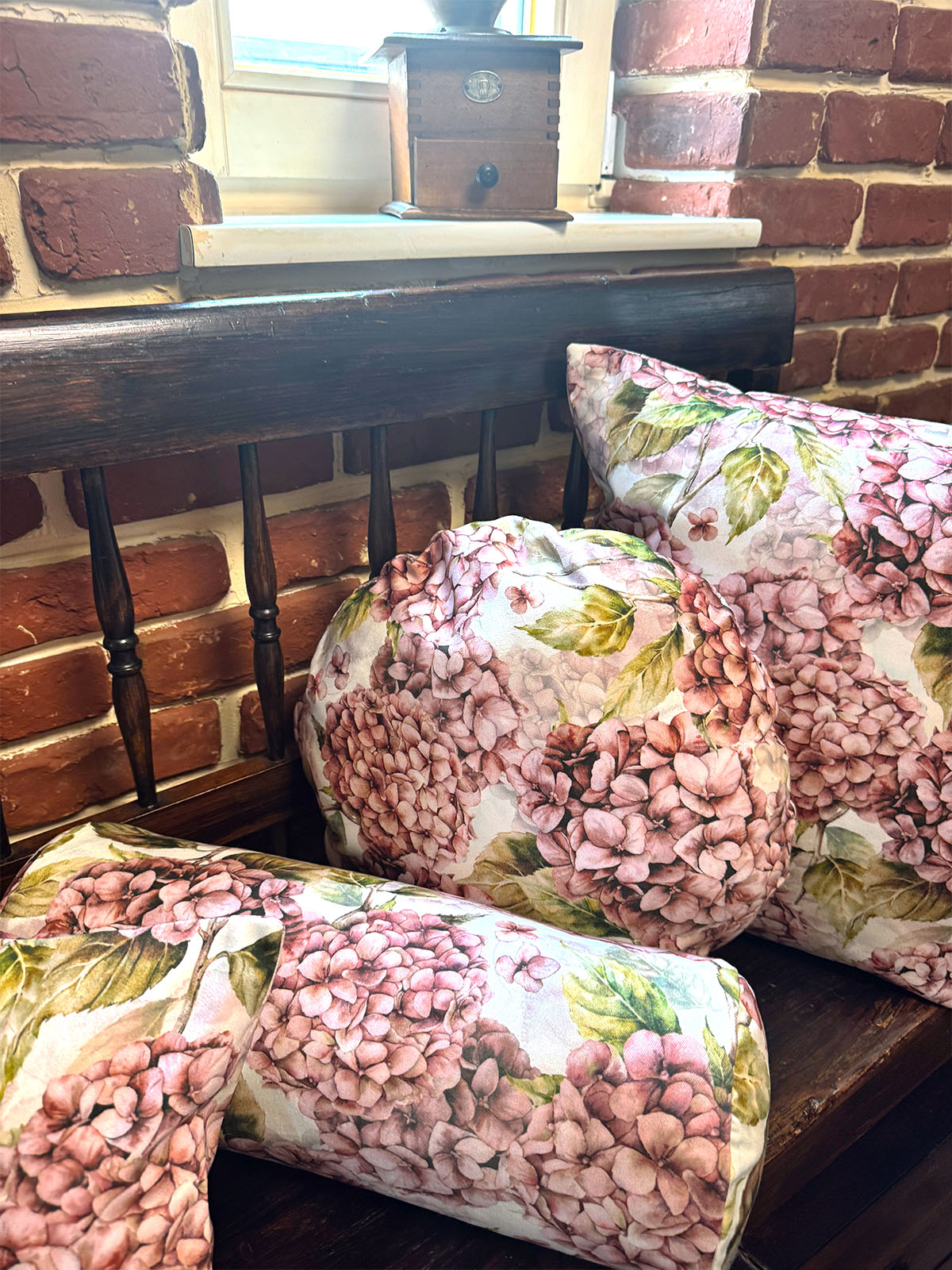Auf einer dunklen Holzbank sind Kissenbezuege im Landhausstil, bedruckt mit rosa/pinken Hortensien-Blueten, drapiert.