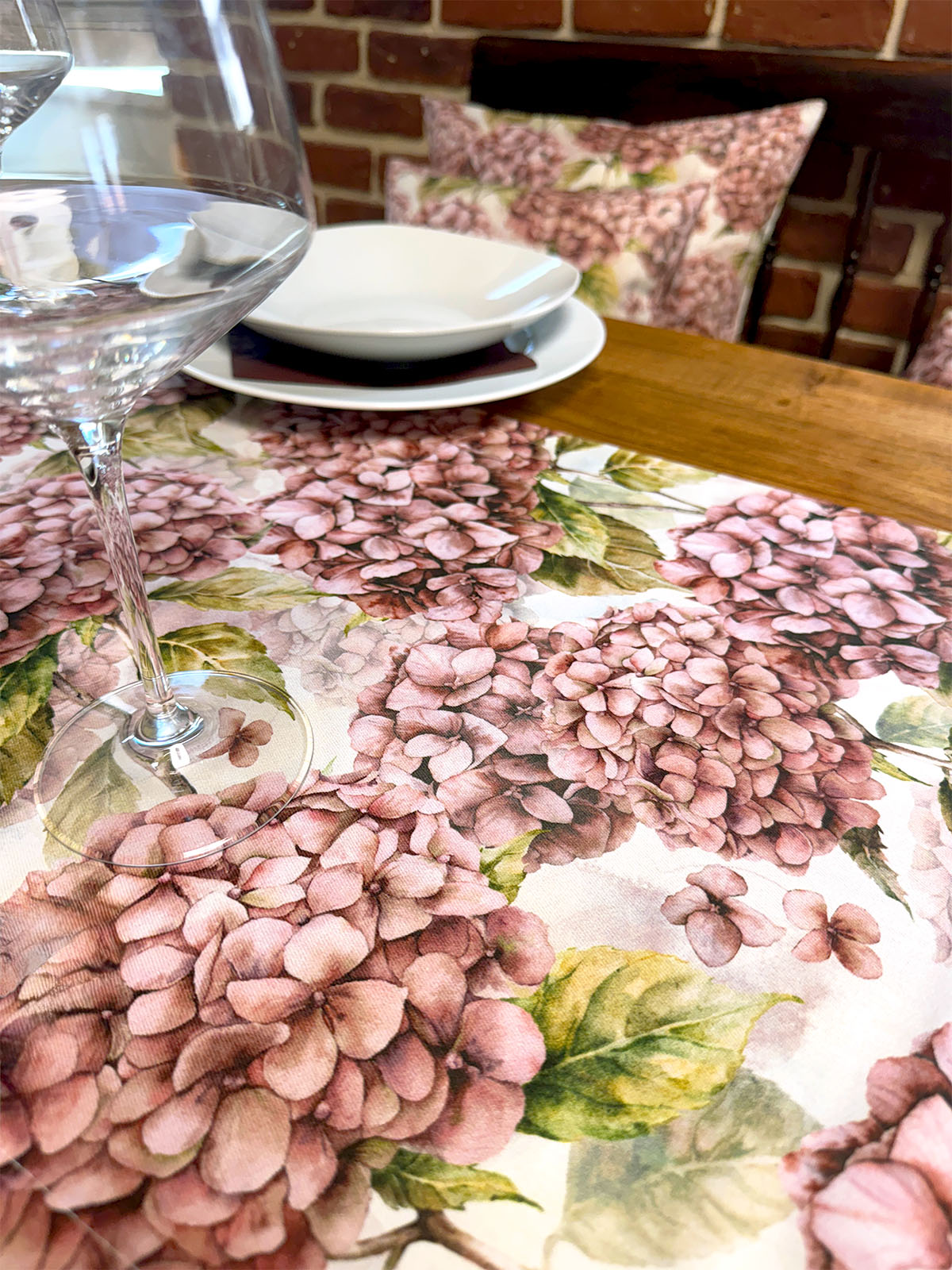 Eine helle, geblümte Tischdecke bedruckt mit rosa/pinken Hortensien auf einem gedeckten Holztisch.