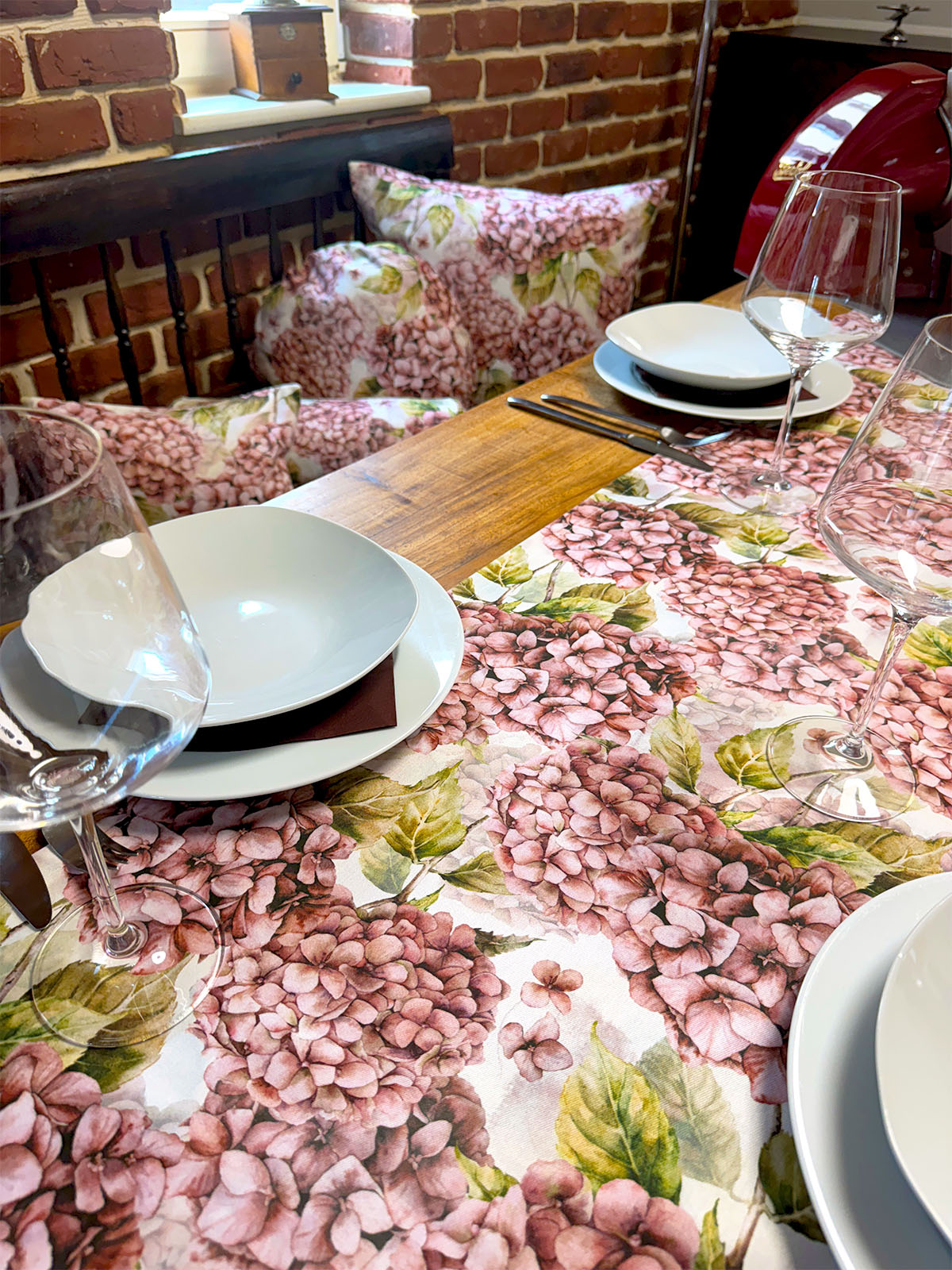 Eine helle, geblümte Tischdecke bedruckt mit rosa/pinken Hortensien auf einem gedeckten Holztisch.