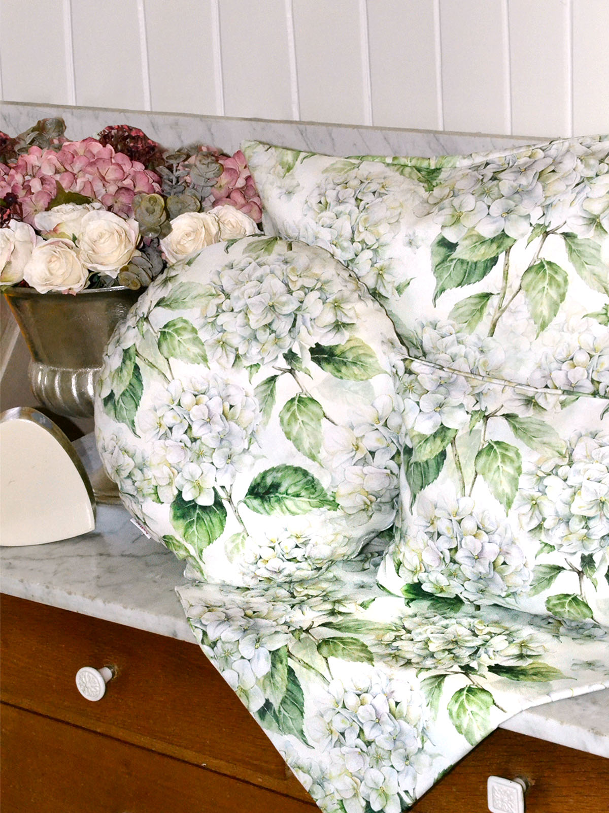 Drei Kissen bezogen mit je einem Kissenbezug im Landhausstil und eine Mitteldecke, bedruckt mit weissen Hortensien. Dekoriert auf einem Sideboard.