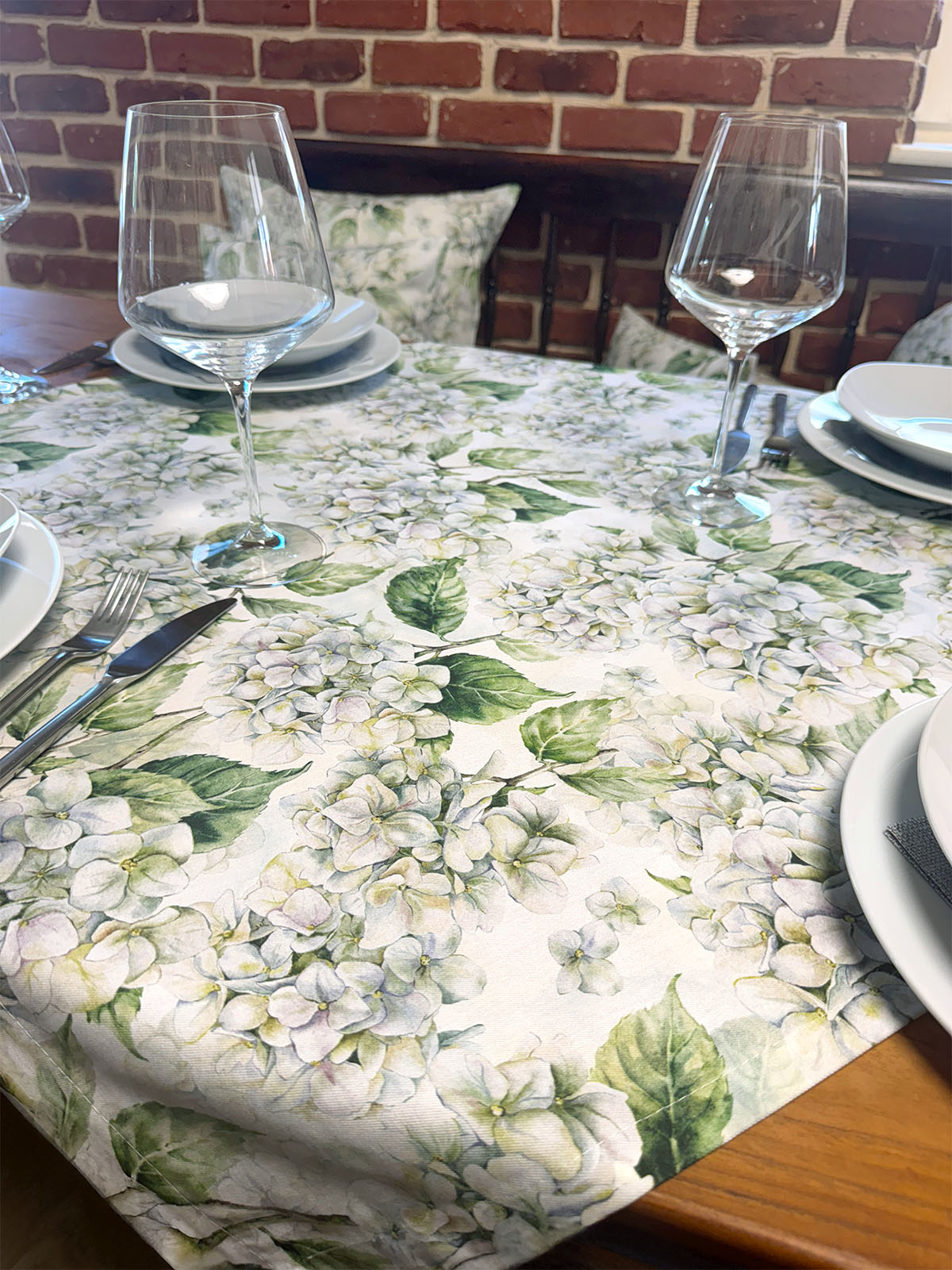 Eine weisse, geblümte Tischdecke bedruckt mit weissen Hortensien auf einem gedeckten Holztisch.