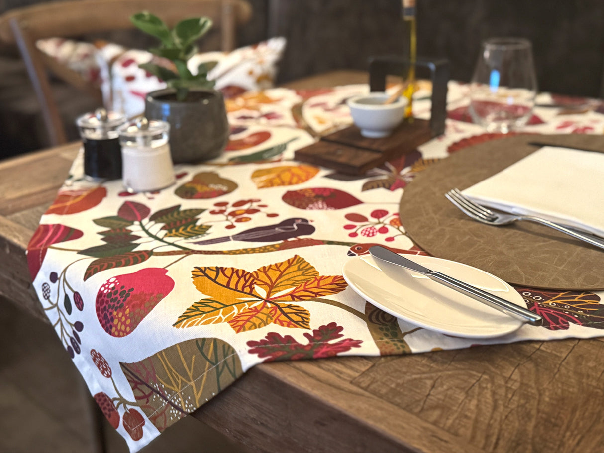 Ein eingedeckter Holztisch mit einer, mit bunten Blaettern, Zweigen und Voegeln bedruckten, Mitteldecke.