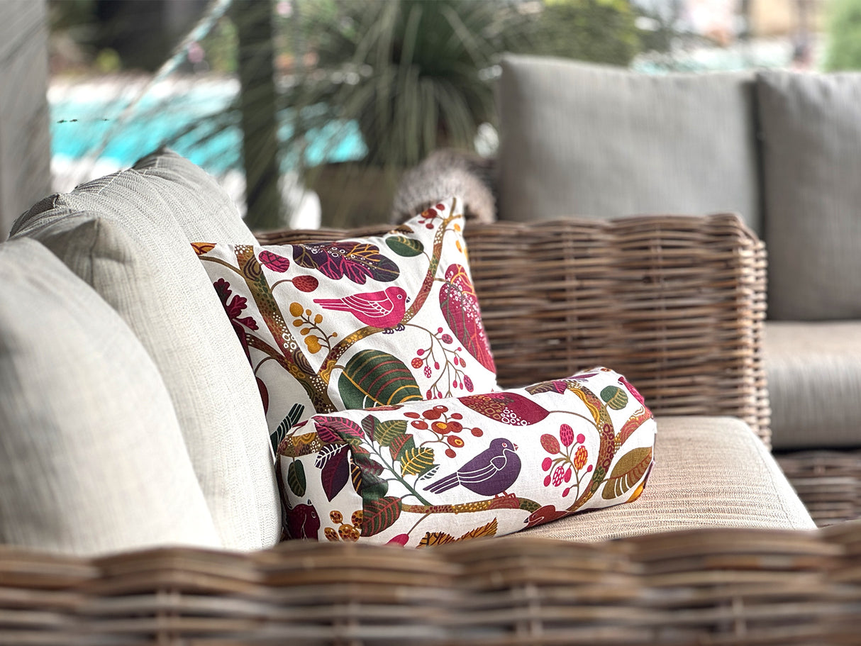 Ein quadratisches Kissen und eine Nackenrolle sind auf einem hellen Sofa drapiert. Sie sind beide mit Kissenbezuegen bezogen die mit bunten Blaettern, Zweigen und Voegeln bedruckt sind.