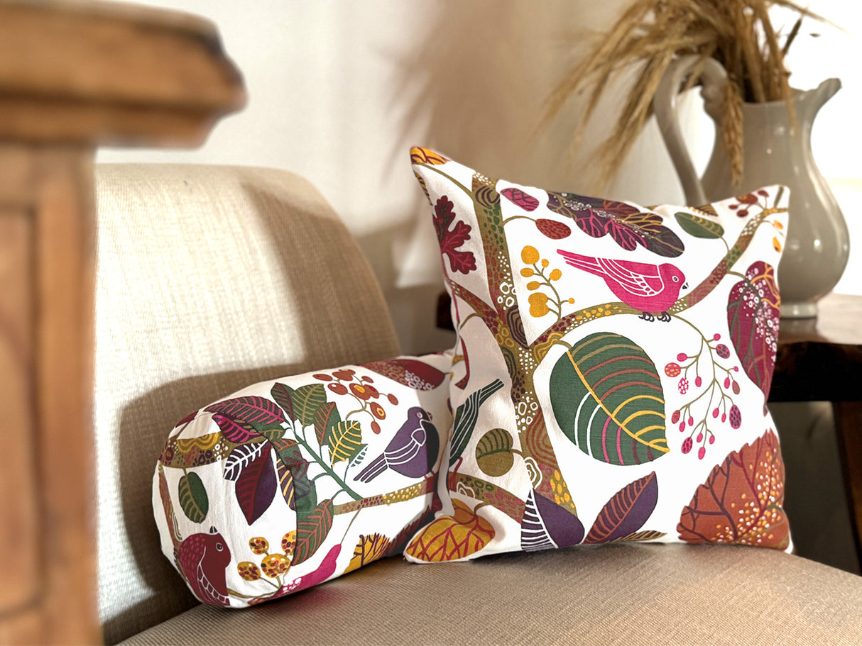 Ein quadratisches Kissen und eine Nackenrolle sind auf einem Sessel drapiert. Sie sind beide mit Kissenbezuegen bezogen die mit bunten Blaettern, Zweigen und Voegeln bedruckt sind.