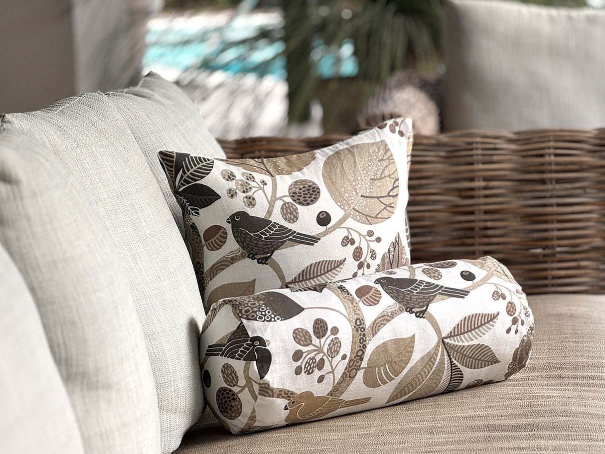Ein quadratisches Kissen und eine Nackenrolle sind auf einem hellen Sofa drapiert. Sie sind beide mit Kissenbezuegen bezogen die mit Blaettern, Zweigen und Voegeln in den Farben taupe und braun bedruckt sind.