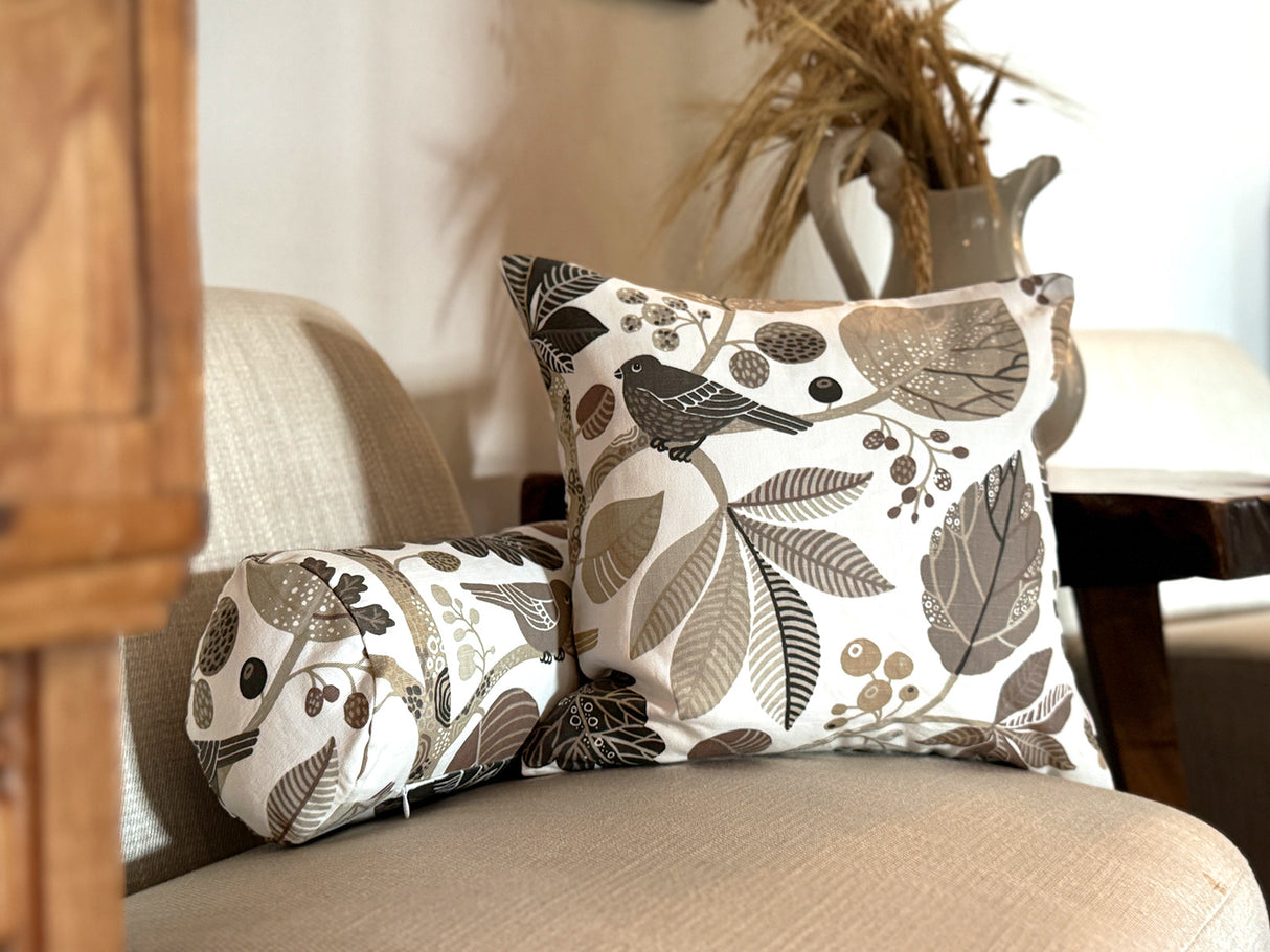 Ein quadratisches Kissen und eine Nackenrolle sind auf einem Sessel drapiert. Sie sind beide mit Kissenbezuegen bezogen die mit Blaettern, Zweigen und Voegeln in den Farben taupe und braun bedruckt sind.