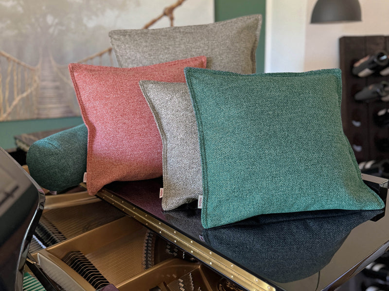 Vier quadratische Kissen und eine Nackenrolle, bezogen mit Boucle Kissenbezuegen in den Farben Petrol, Grau und Rot auf einem Klavier dekoriert.