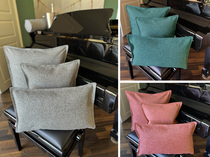 Drei Kissen, in unterschiedlichen Groessen, auf einem Klavierhocker vor einem Klavier. Bezogen mit Boucle Kissenhuellen in den Farben Grau, Petrol und Rot.