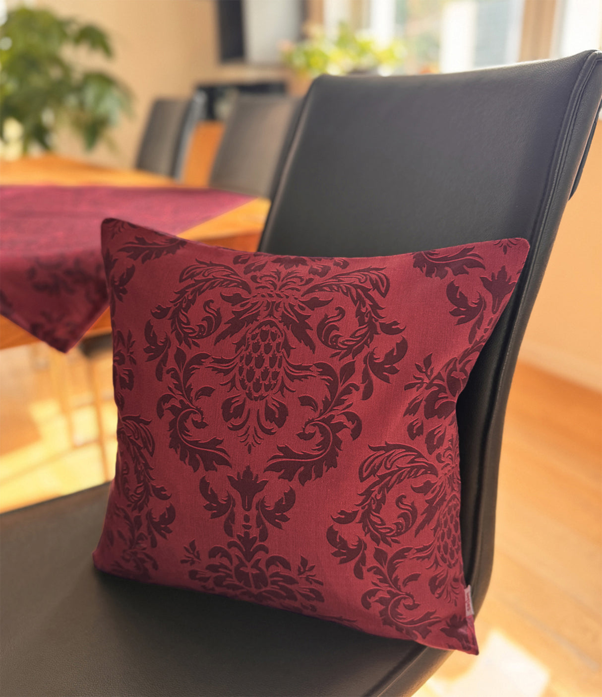 Auf einem schwarzen Lederstuhl, lehnt ein Kissen, bezogen mit einem roten Kissenbezug, der mit barocken Ornamenten in einem dunkleren Roten, bedruckt ist.