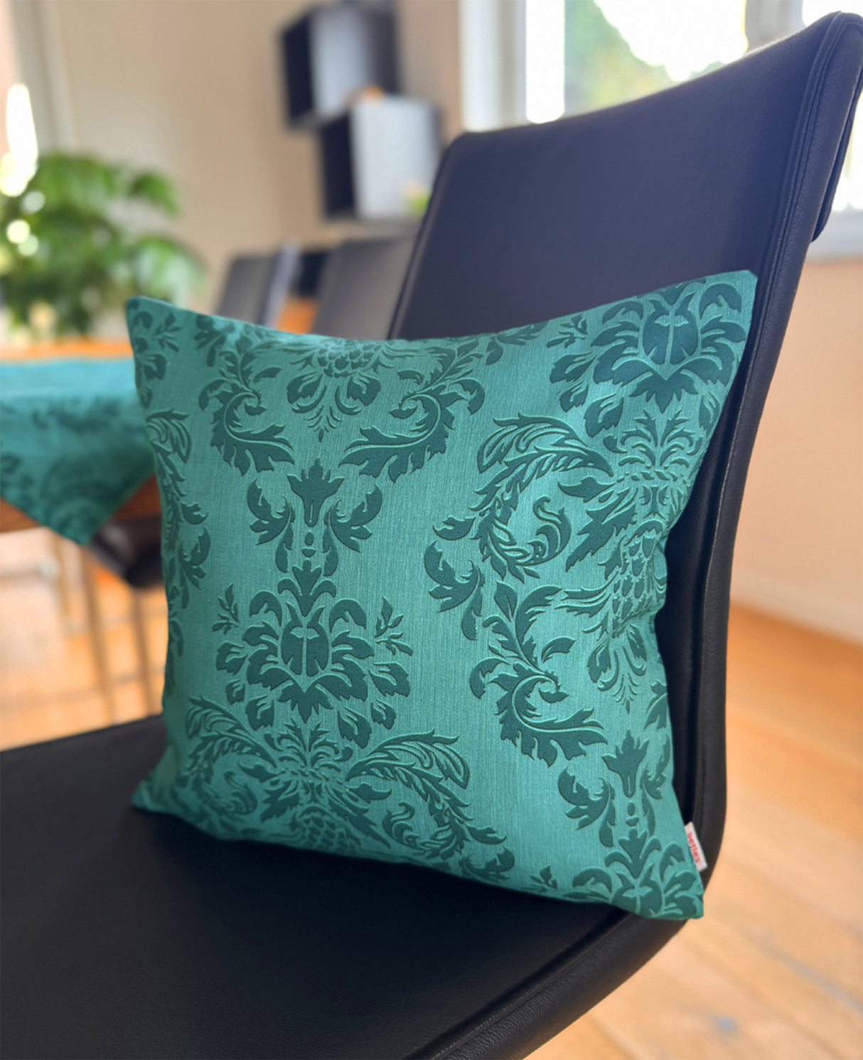 Auf einem schwarzen Lederstuhl, lehnt ein Kissen, bezogen mit einem petrolfarbenen Kissenbezug, der mit barocken Ornamenten in einem dunkleren Petrol, bedruckt ist.