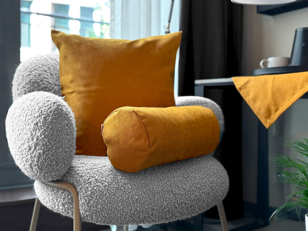 Ein quadratisches Kissen und eine Nackenrolle, drapiert auf einem Sessel, bezogen mit unifarbenen Kissenbezuegen in der Farbe curry-gelb.