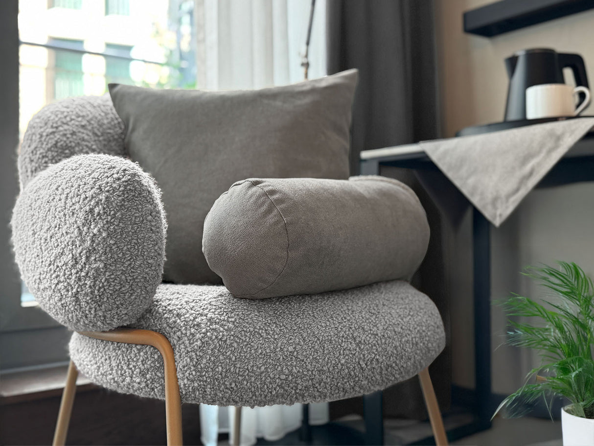 Auf einem Sessel ist ein quadratisches Kissen und eine Nackerolle in der Farbe greig-taupe drapiert. Im Hintergrund liegt auf einem Tisch eine Mitteldecke in der gleichen Farbe.