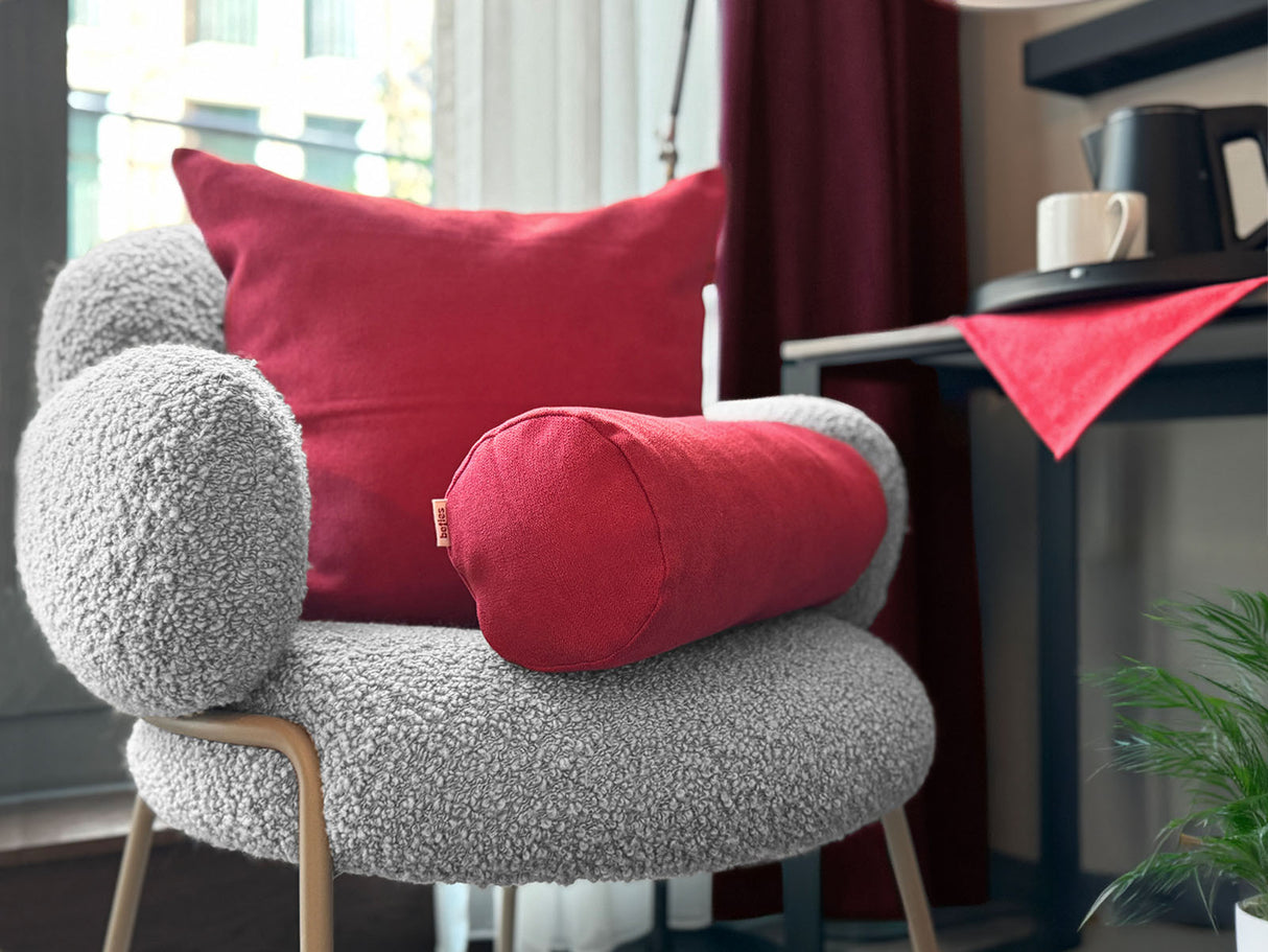 Auf einem Sessel ist ein quadratisches Kissen und eine Nackerolle in der Farbe himbeer-rot drapiert. Im Hintergrund liegt auf einem Tisch eine Mitteldecke in der gleichen Farbe.