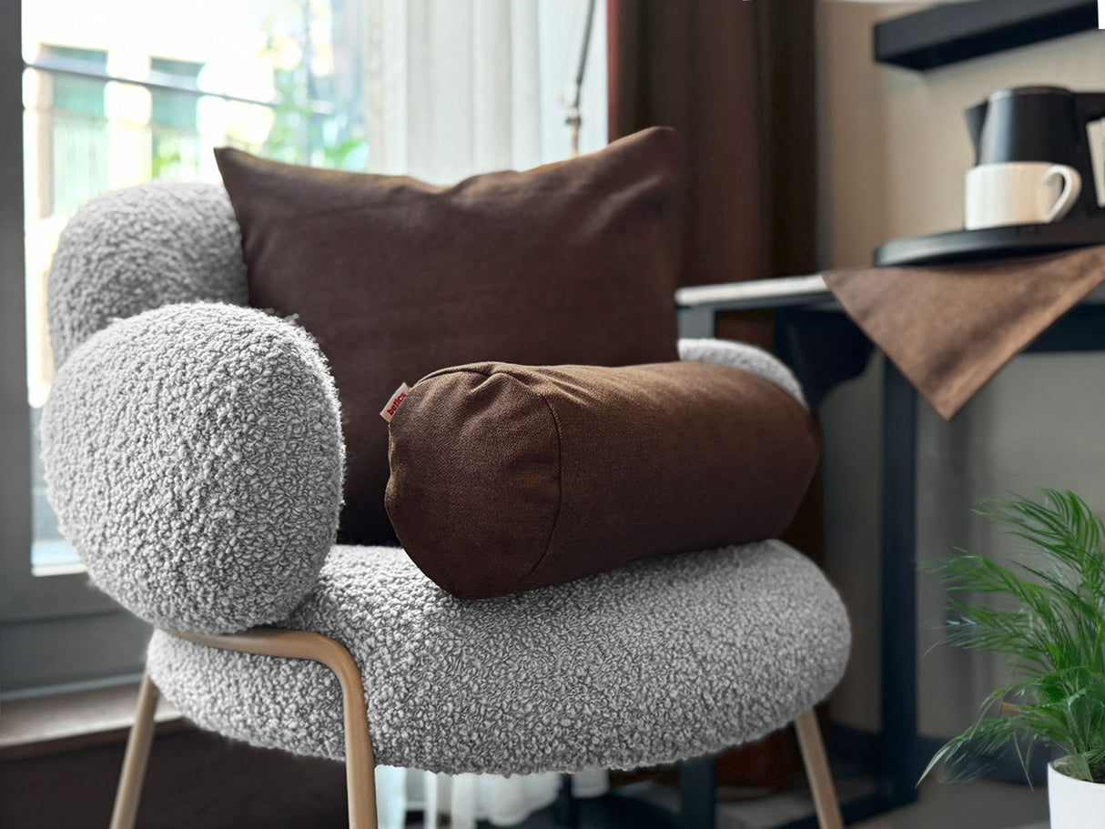 Auf einem Sessel ist ein quadratisches Kissen und eine Nackenrolle in der Farbe kaffeebohne-braun drapiert. Im Hintergrund liegt auf einem Tisch eine Mitteldecke in der gleichen Farbe.